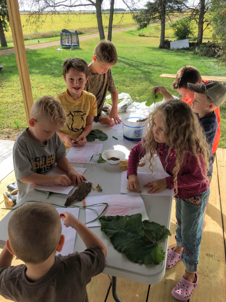 Kids around a long rectangle table on a porch outside making leaf art.